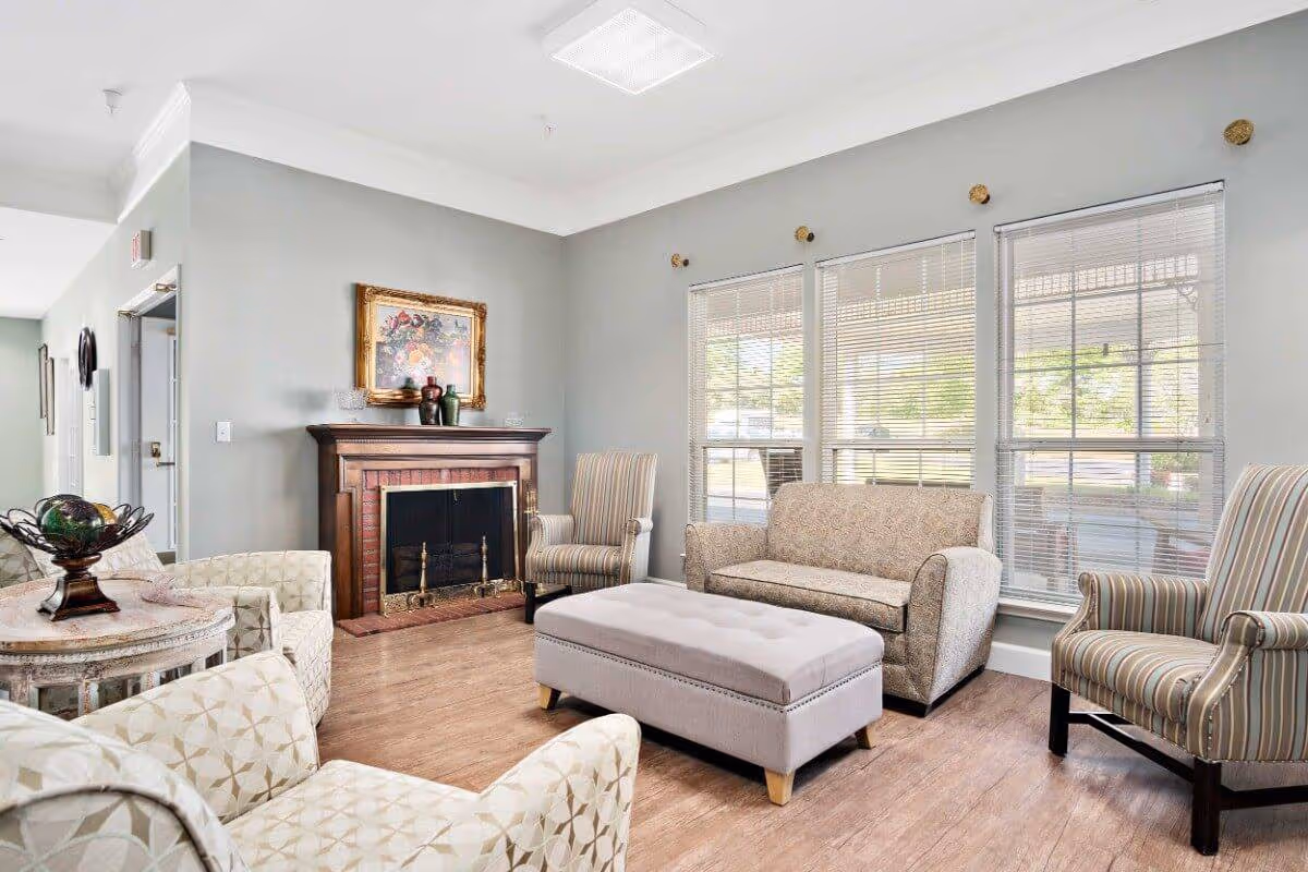 A bright and cozy living room with light gray walls and wood flooring. The room features a patterned sofa, two striped armchairs, two patterned armchairs, and a large upholstered ottoman in the center. There is a wooden fireplace with a brick interior and a framed floral painting above it. Large windows with blinds allow natural light to fill the space.