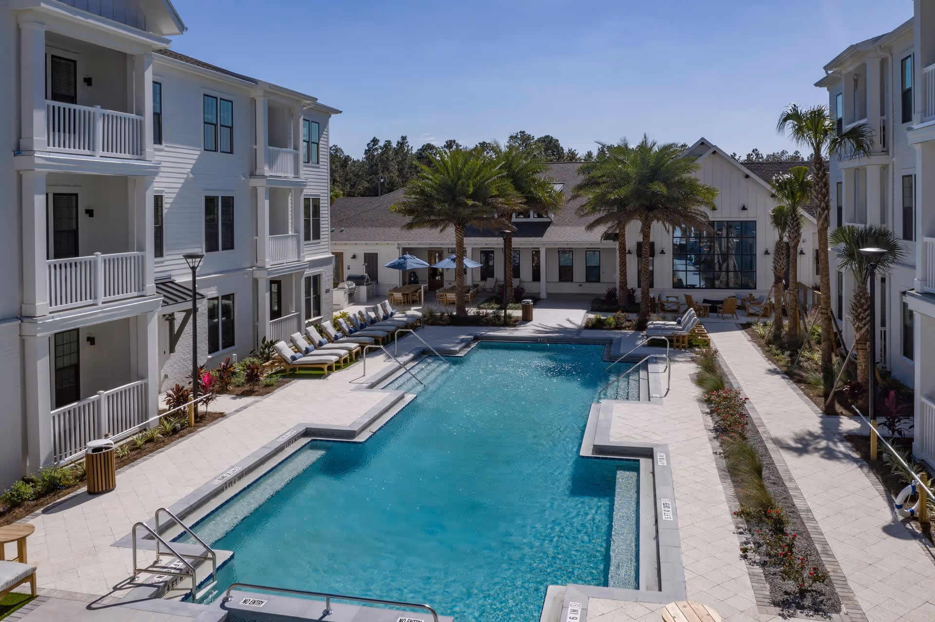 Courtyard swimming pool flanked by white three-story residential buildings, palm trees, and lounge chairs.