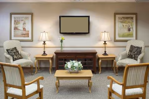 Senior living facility lounge with seating area, coffee table, TV, lamps, and framed art.