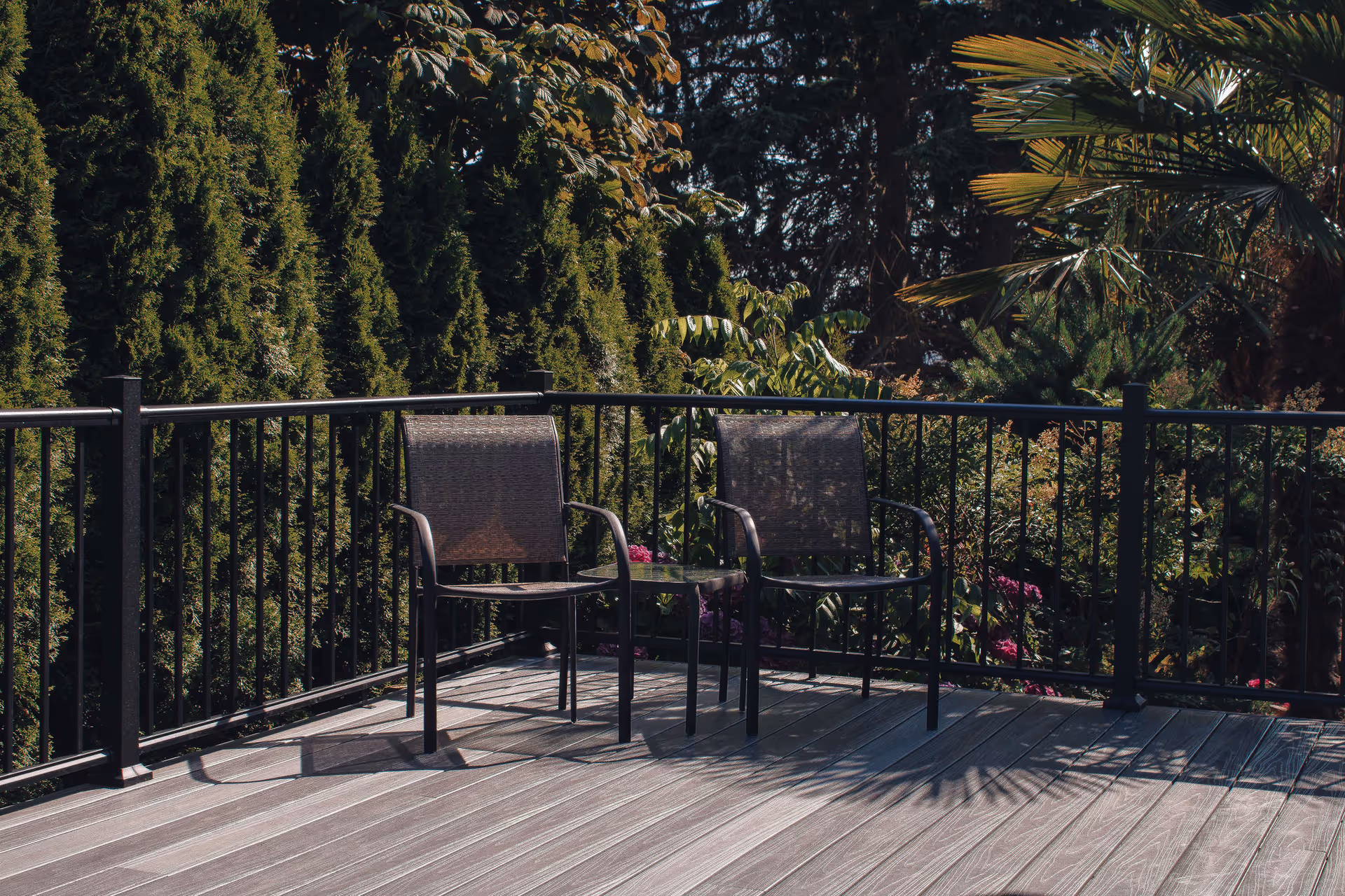 Two outdoor chairs and a small table sit on a wooden deck with a black railing in front of dense trees and plants.
