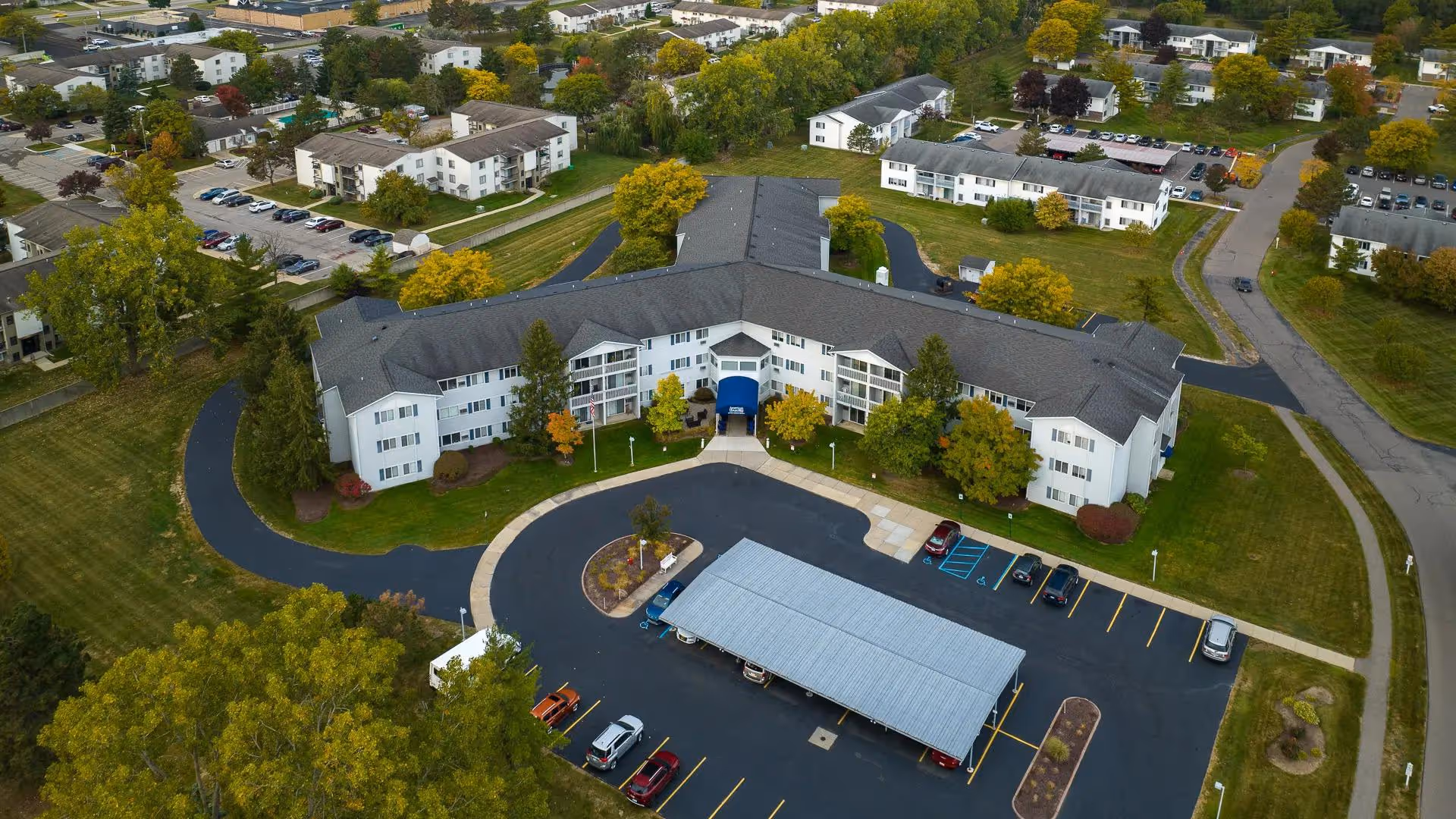 Aerial view of a curved, three-story senior living building with a central entrance, parking lot with a covered carport, and surrounding lawns and trees.