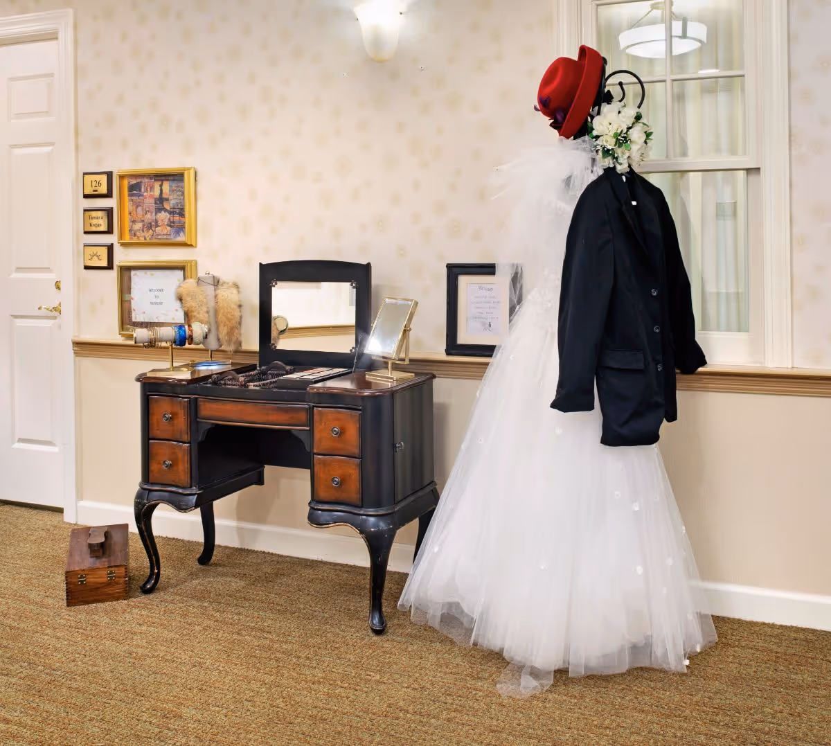An interior room with a vintage wooden vanity table holding jewelry and a mirror. Next to it is a mannequin dressed in a white wedding gown with a black jacket and a red hat, adorned with white flowers. The wall behind has framed pictures and a door labeled 126 Tamara Kogan.