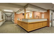 Interior view of a nursing facility hallway with a wooden reception desk in the center. The walls are painted in neutral tones with some peach accents, and the floor is carpeted. The hallway extends into the distance with doors and framed pictures along the walls.