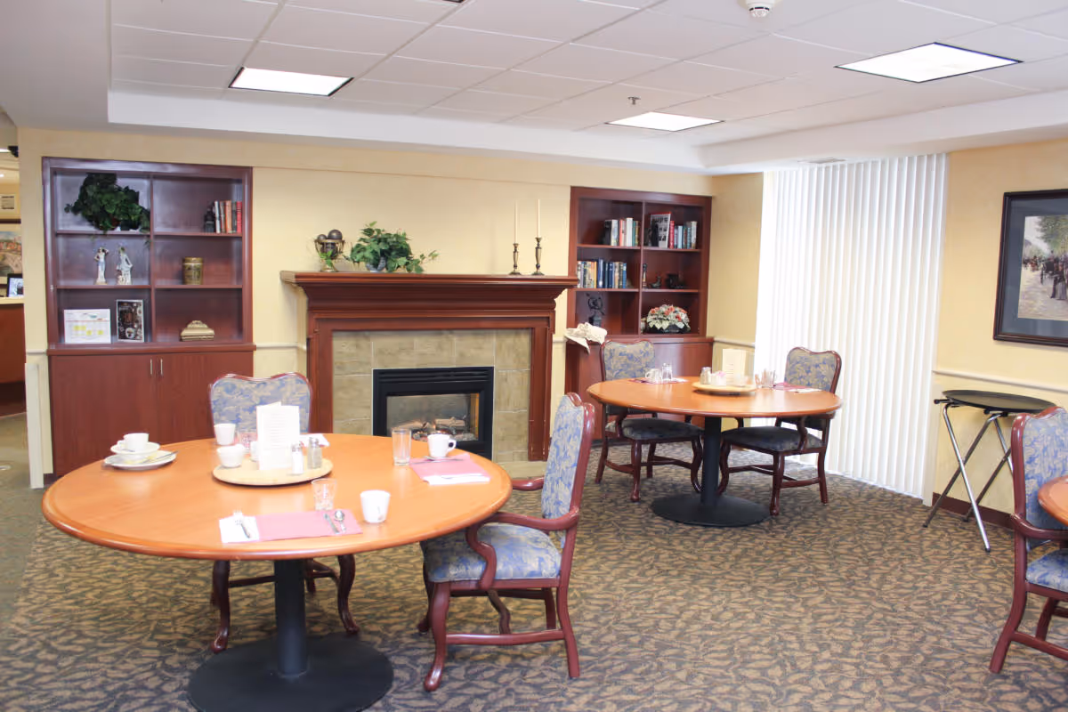 A cozy dining area in an assisted living facility with round wooden tables set with cups, glasses, and napkins. The room features a fireplace with a wooden mantel, built-in bookshelves with books and decorative items, patterned carpet, and vertical blinds covering a window.