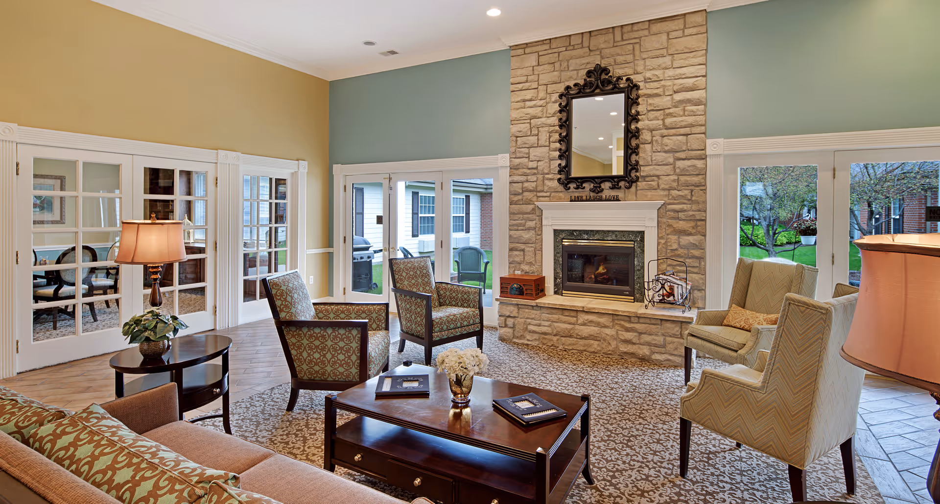 A cozy senior living facility common area with a stone fireplace, a decorative mirror above it, and several upholstered chairs and a sofa arranged around a wooden coffee table. The room has large glass doors leading outside, patterned carpet, and warm lighting from table lamps.