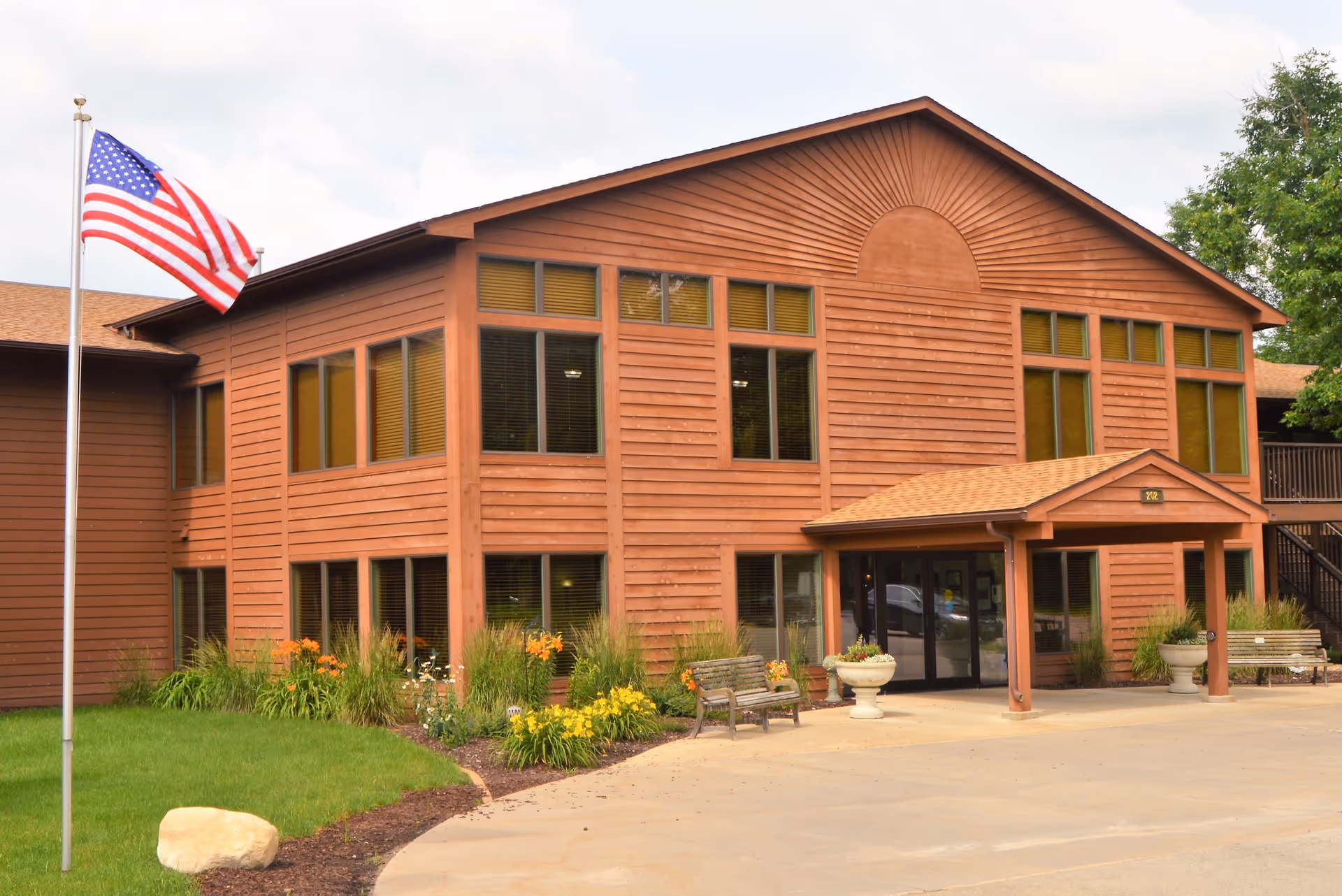 Exterior view of a two-story brown wooden building with multiple windows, a covered entrance, benches, flower beds, and an American flag on a flagpole in front.