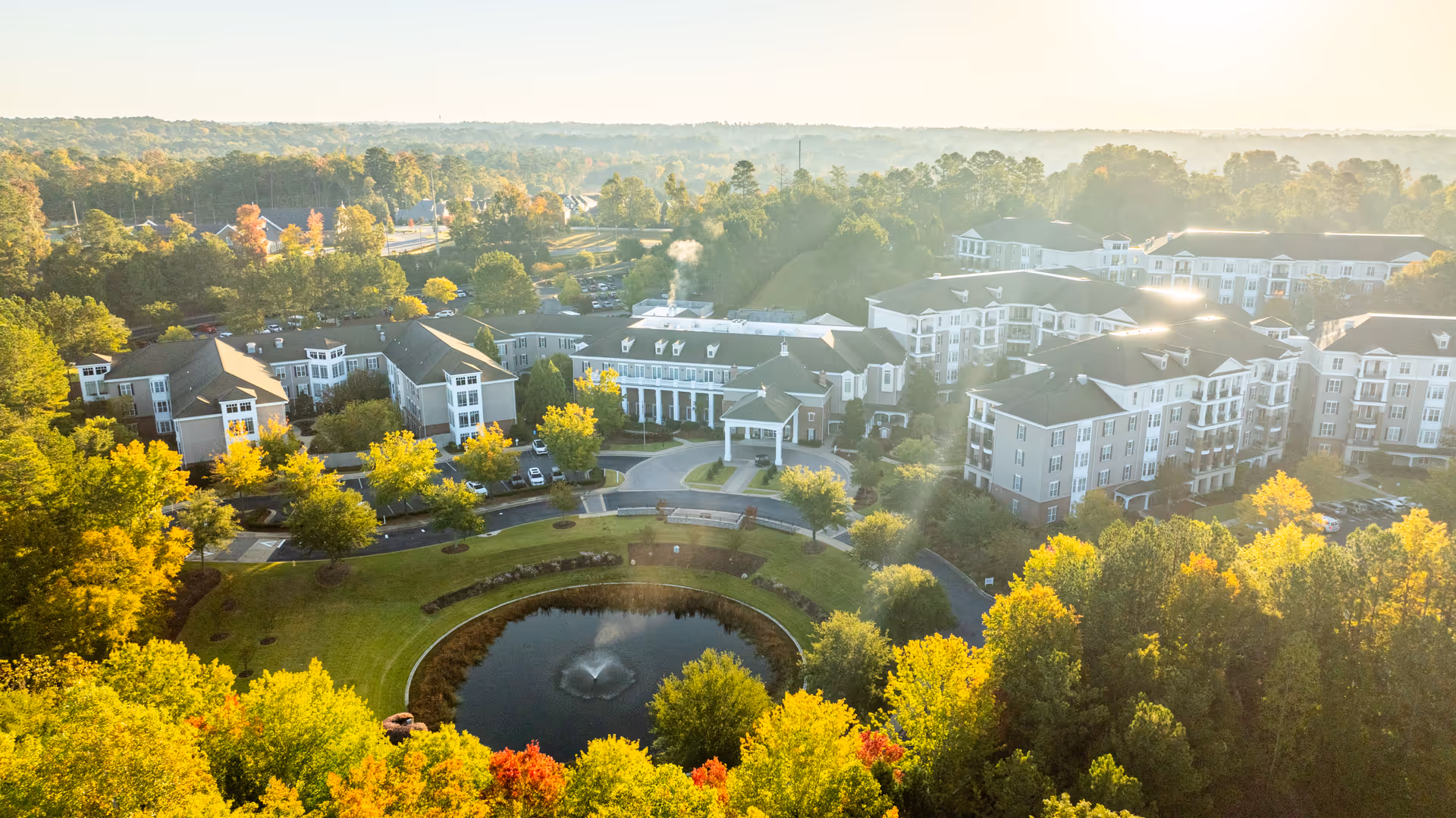 Aerial view of Spring Harbor at Green Island senior living facility surrounded by trees with autumn foliage. The complex includes multiple multi-story buildings arranged around a circular driveway and a pond with a fountain in the center. The scene is bathed in soft sunlight with a misty background.