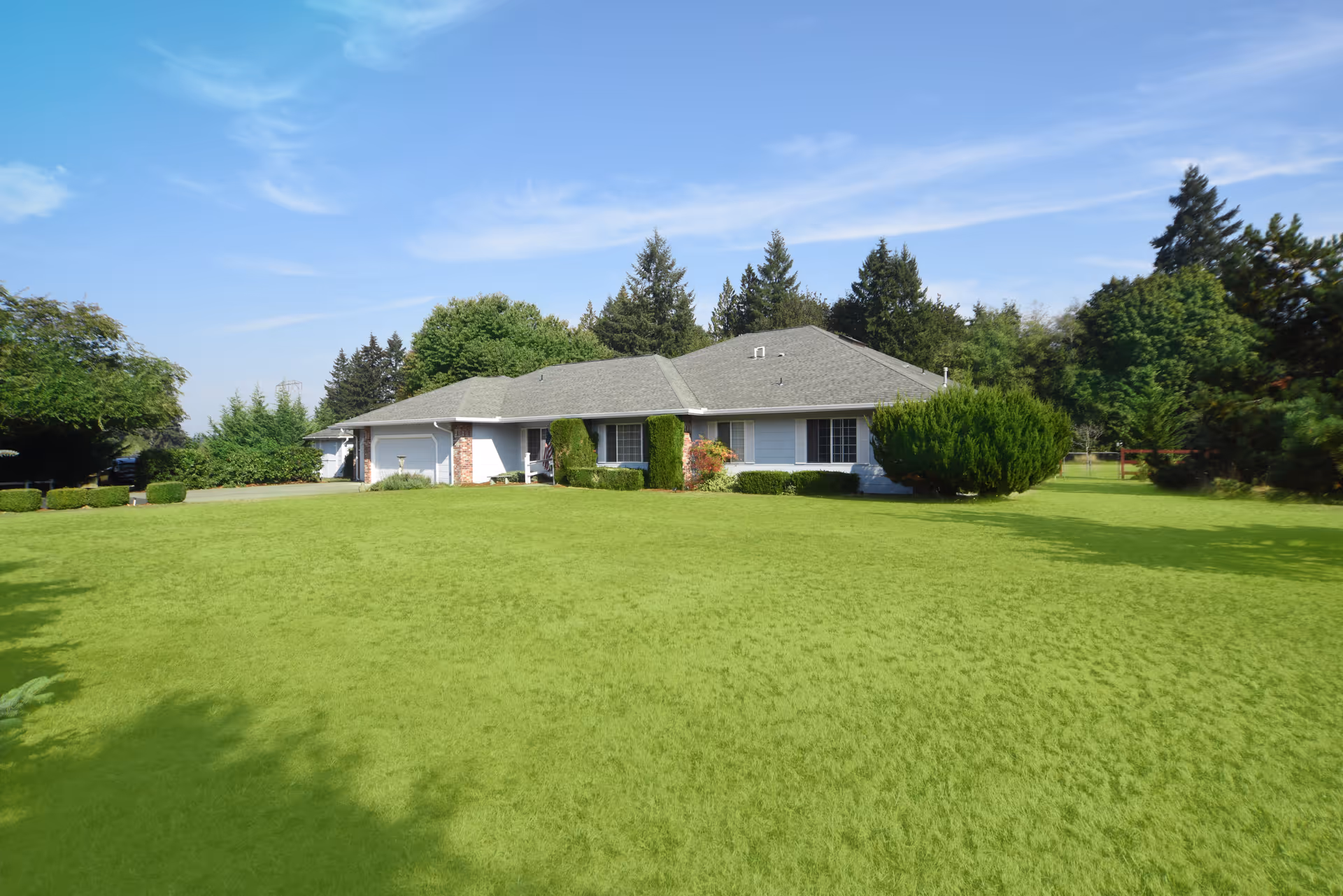 Single-story residential building with a gray roof and white exterior walls, surrounded by a large green lawn and various trees under a blue sky with some clouds.
