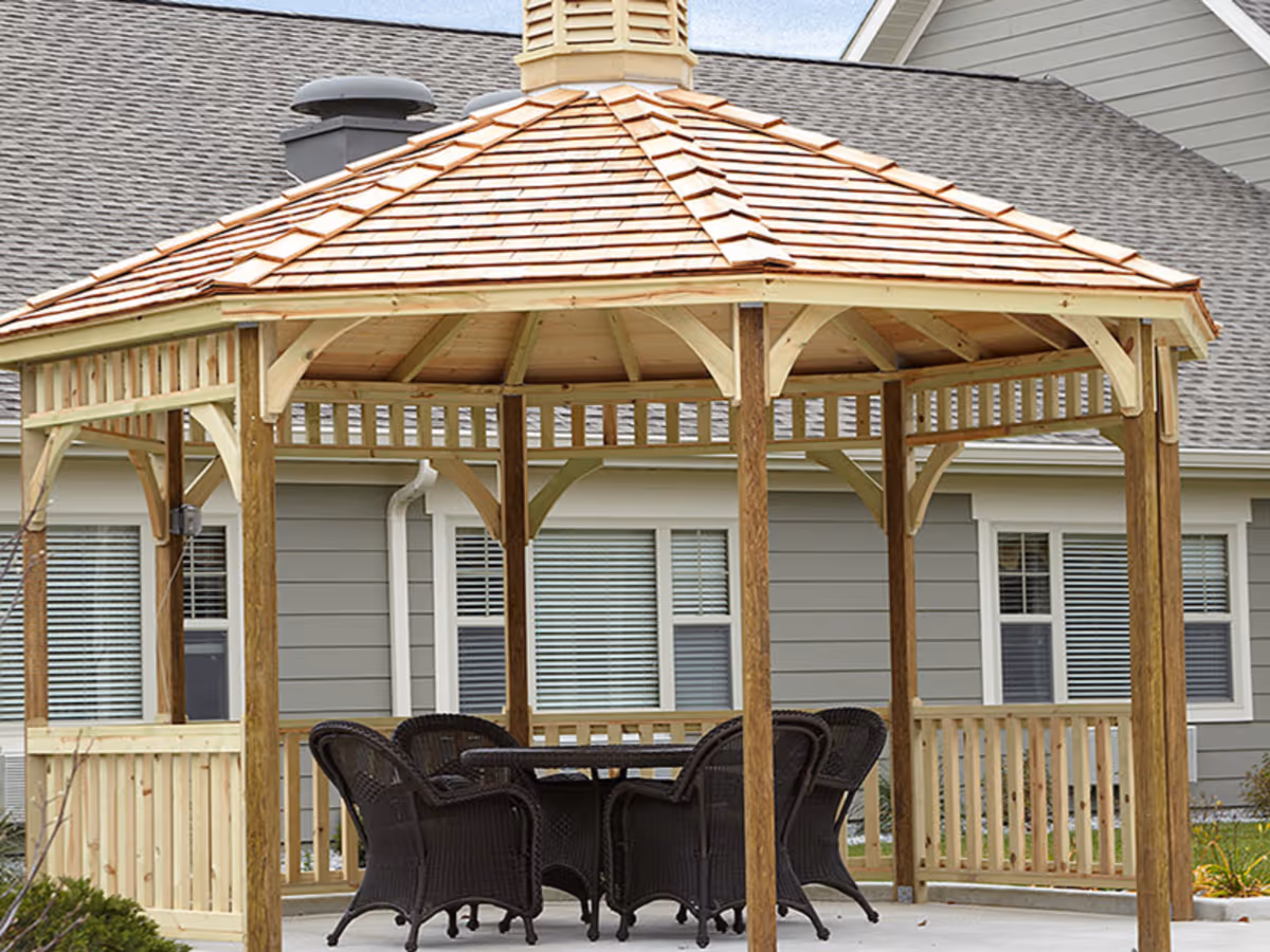 A wooden gazebo with a shingled roof and open sides, containing a round table surrounded by six dark wicker chairs, situated outdoors in front of a gray building with white-trimmed windows.