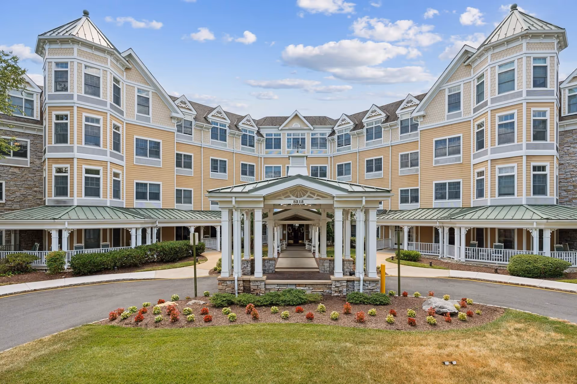 Front exterior view of a large senior living facility building with yellow siding, multiple windows, and a covered entrance with white columns. There is a circular driveway and landscaped flower beds in front under a partly cloudy sky.