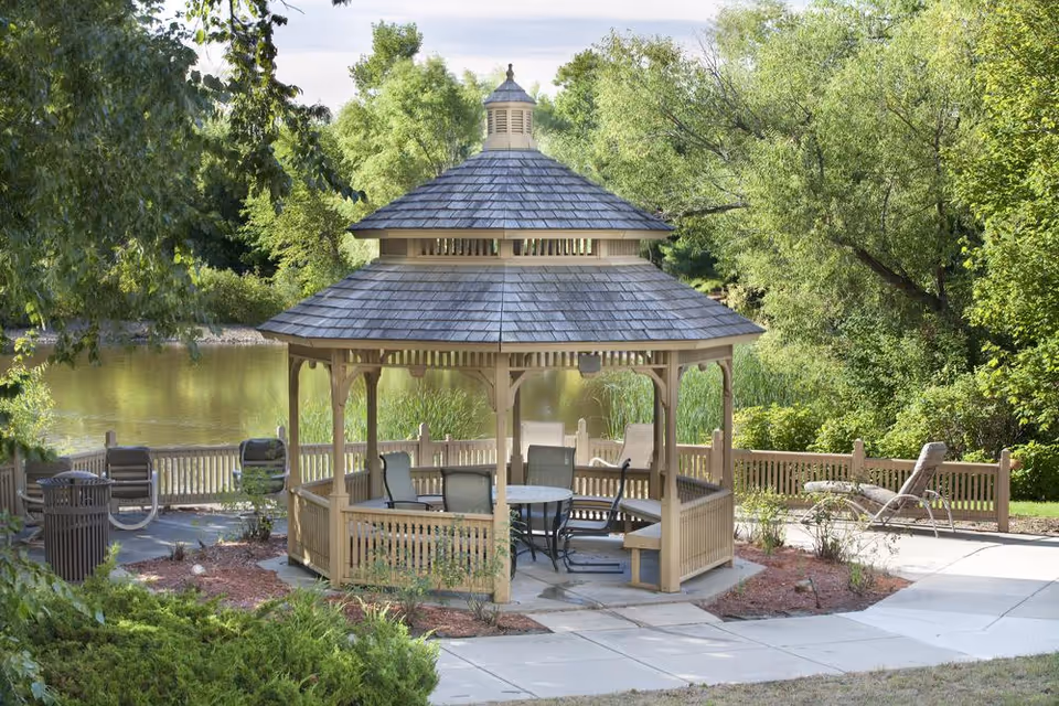 A wooden gazebo with a shingled roof situated in a landscaped outdoor area near a pond, surrounded by green trees and bushes. The gazebo contains a round table and several chairs. Nearby, there are lounge chairs and a trash can along a paved walkway.