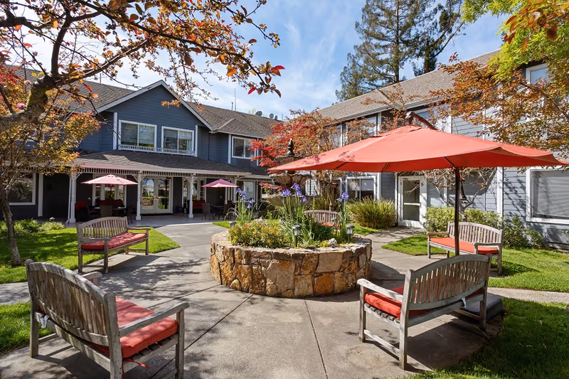 Courtyard with wooden benches and red umbrellas surrounding a central stone planter in front of a two-story gray residential building.