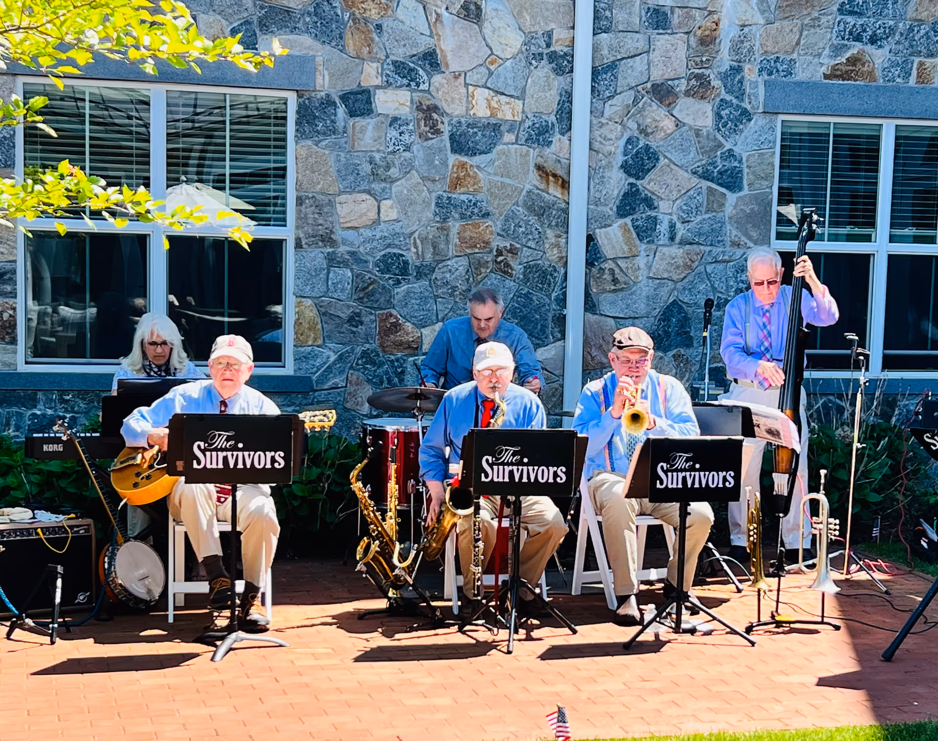 A group of six elderly musicians performing outdoors in front of a stone wall with windows. They are seated and playing various instruments including guitar, saxophone, trumpet, drums, and double bass. Music stands in front of them display the name 'The Survivors'.