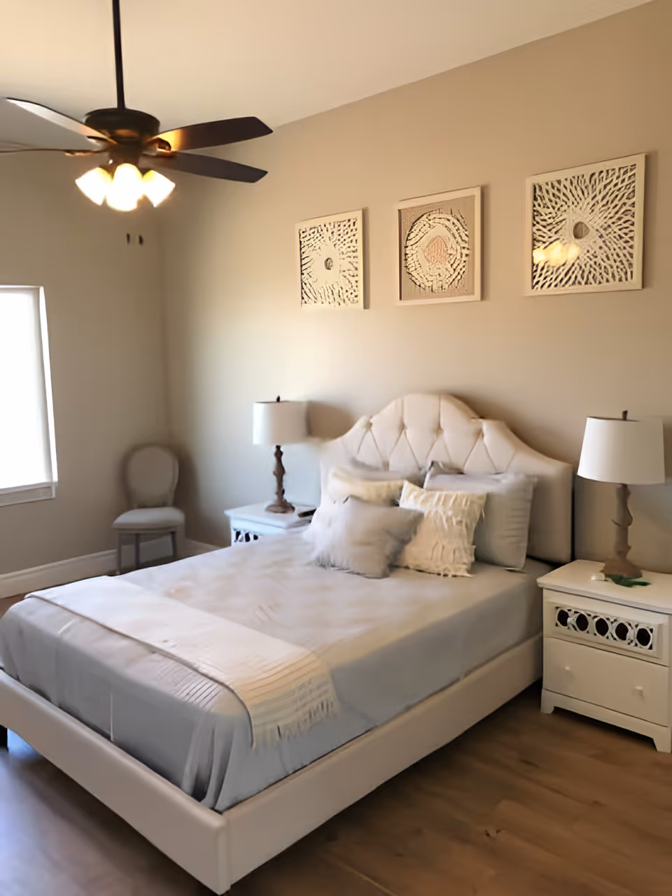 Light-filled bedroom with a tufted bed, two nightstands and lamps, wall art, and a ceiling fan.