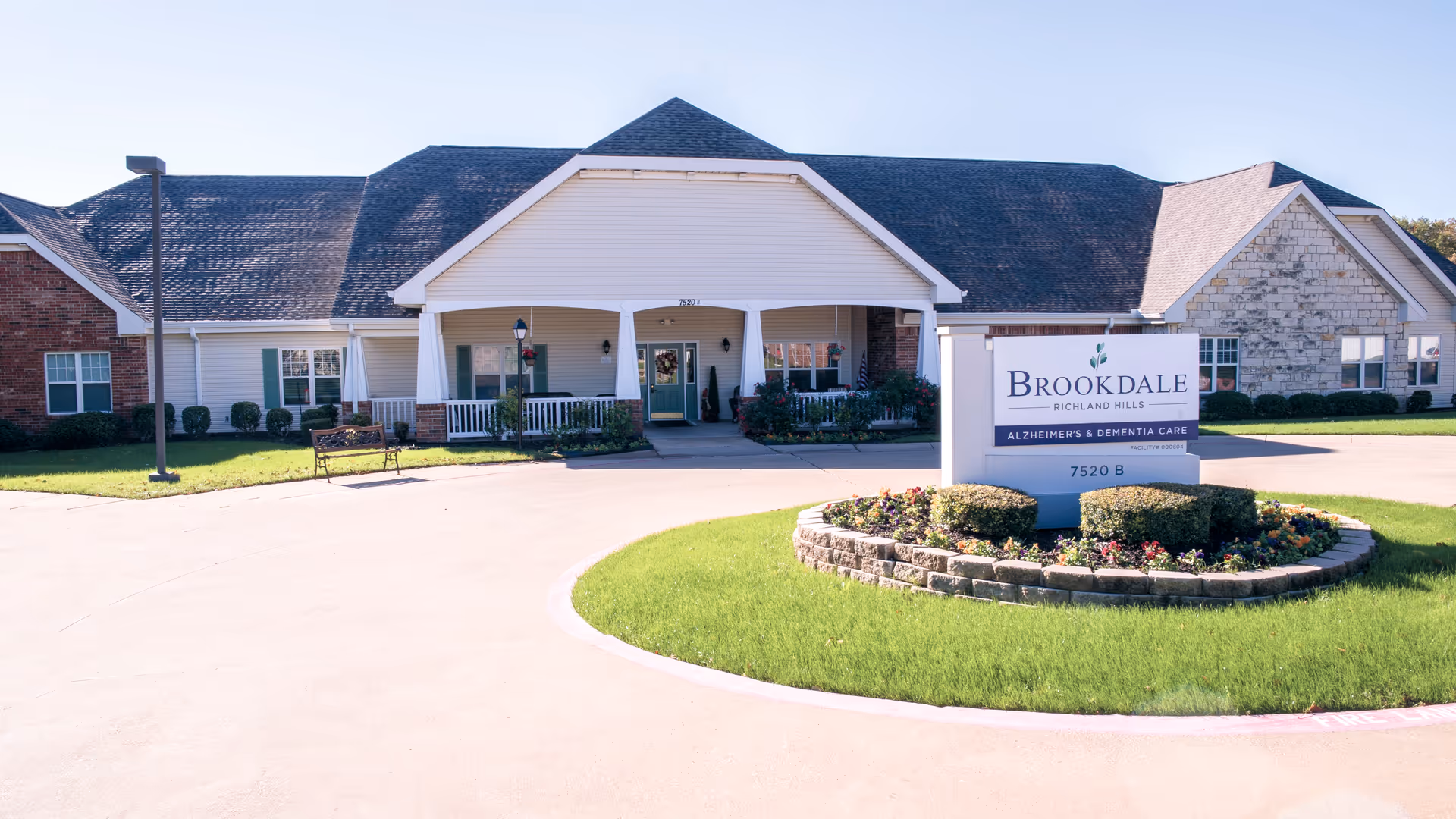 Exterior view of Brookdale Richland Hills, a single-story building with a large covered entrance, white siding, brick and stone accents, surrounded by a well-maintained lawn and a circular flower bed with a sign displaying the facility name and services.