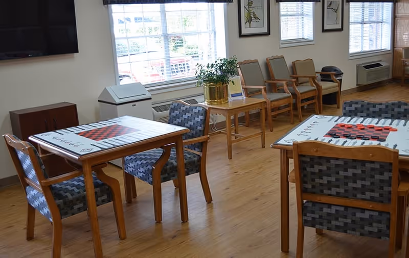 A bright room with wooden flooring featuring two square tables with checkerboard game designs on the tabletops. Each table is surrounded by cushioned chairs with patterned upholstery. Along the wall, there are several wooden chairs with armrests, a small wooden table with a potted plant, and large windows letting in natural light. A flat-screen TV is mounted on the wall.