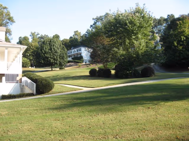 A grassy outdoor area with a paved walkway winding through it, surrounded by trees and bushes. Two white buildings are partially visible on the left and in the background, under a clear blue sky.