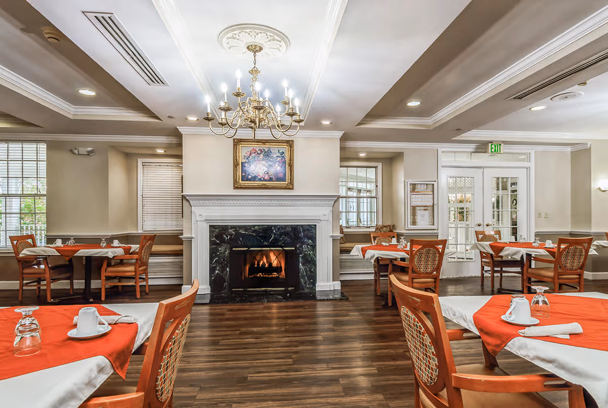 Dining room with wooden floors and multiple tables covered with white and red tablecloths. Each table is set with cups, glasses, and napkins. A lit fireplace with a black marble surround and a framed floral painting above it is centered on the far wall. A chandelier hangs from the ceiling, and there are windows and glass doors in the background.