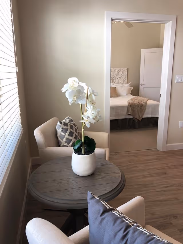 A cozy sitting area with two beige armchairs around a round wooden table holding a white vase with white orchids. In the background, an open doorway reveals a bedroom with a neatly made bed, beige and white bedding, and a closed white door.