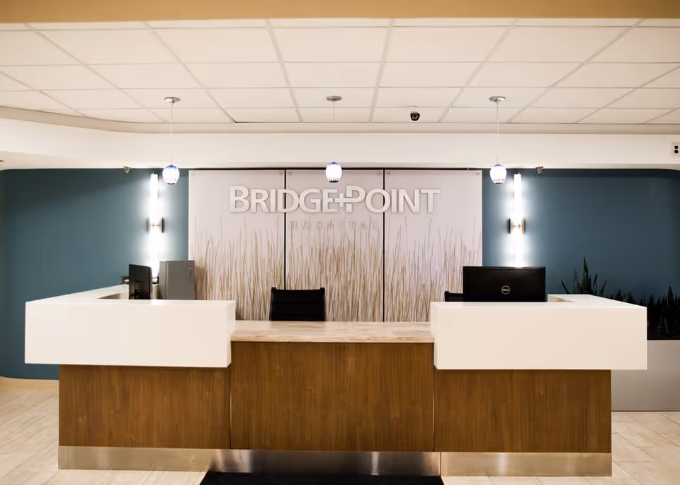 Reception desk area inside BridgePoint Hospital Capitol Hill with a wooden front desk, two computer monitors, three hanging pendant lights, and a backdrop featuring the BridgePoint Hospital logo and a design of tall grass.