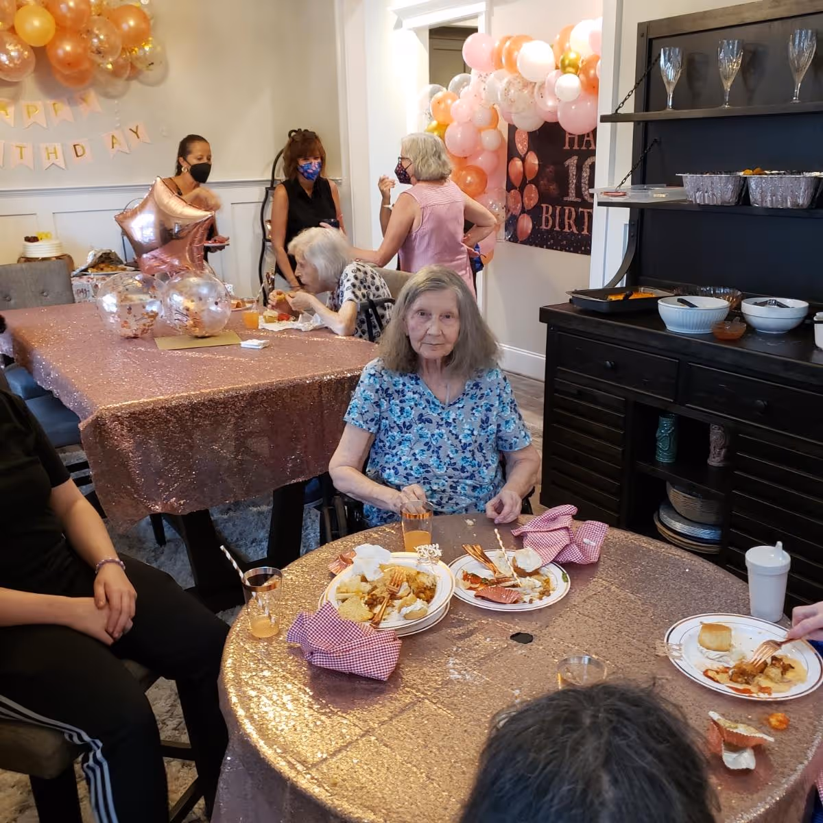 Elderly residents and staff gathered around a decorated dining table with balloons and birthday decorations, eating from plates.