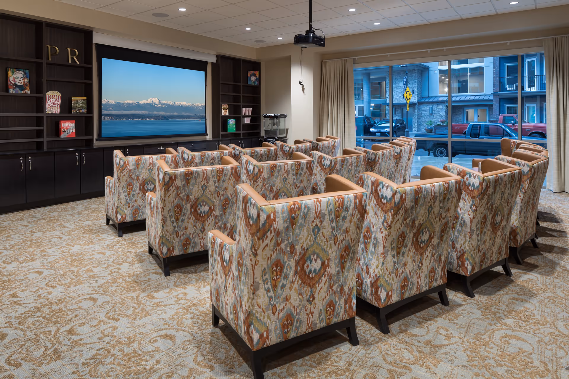 A cozy media room with multiple patterned armchairs arranged in rows facing a large screen displaying a scenic view of mountains and water. The room has built-in dark wood shelving with books and decorative items, a popcorn machine, and large windows with curtains showing a street view outside.