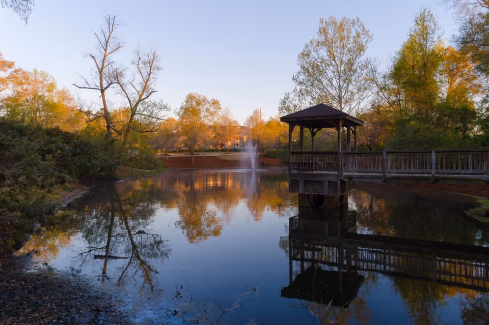 A serene outdoor scene featuring a small pond with a wooden gazebo on stilts connected by a bridge. Trees with autumn foliage surround the pond, and a water fountain sprays in the center of the pond, reflecting the trees and gazebo in the calm water.
