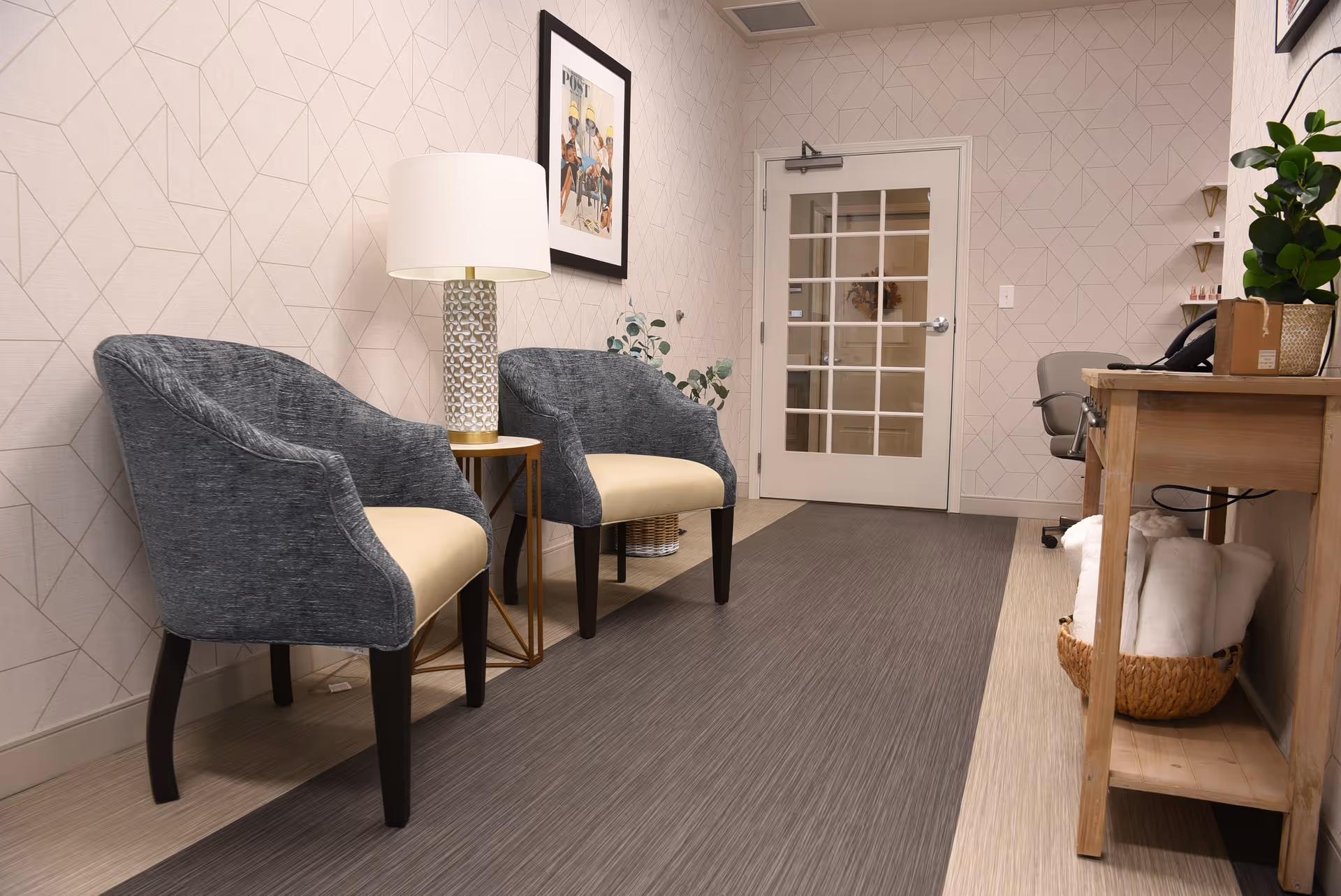 A cozy interior waiting area with two gray upholstered chairs with cream-colored seats, a small round side table with a decorative lamp, a framed picture on the wall, a wooden desk with a plant and office phone, and a basket with rolled white towels underneath the desk. The walls have a subtle geometric pattern, and there is a white door with glass panels at the end of the room.