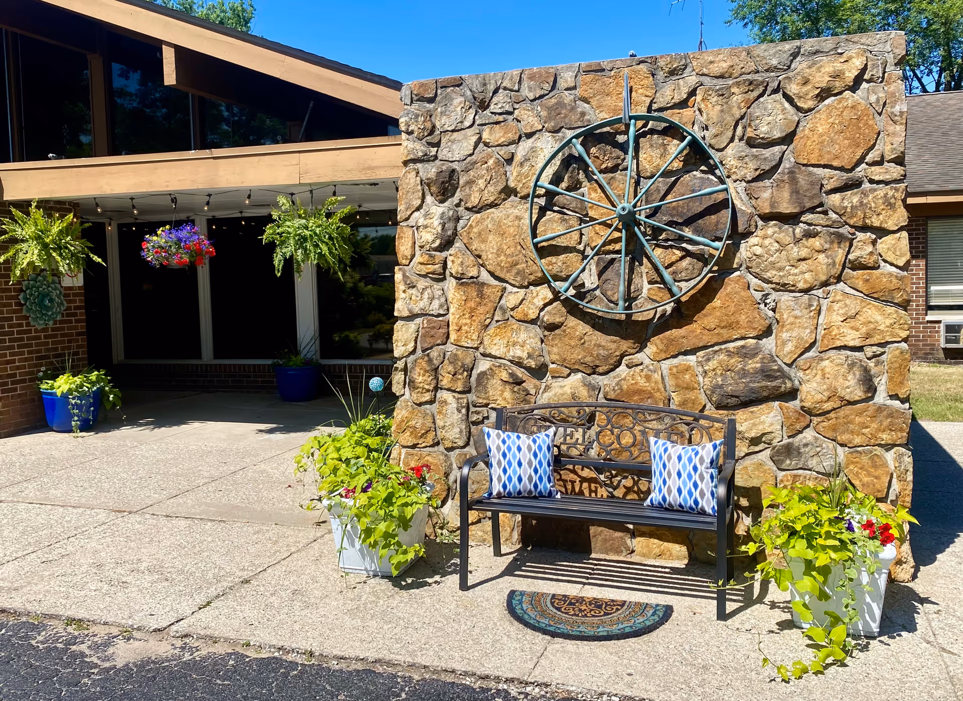 Outdoor seating area at Aperion Care Demotte featuring a stone wall with a decorative metal wagon wheel mounted on it. In front of the wall is a black metal bench with two blue and white patterned cushions. There are potted plants with green foliage and flowers on either side of the bench. The building exterior includes brick walls, large windows, and hanging flower baskets under a wooden overhang.