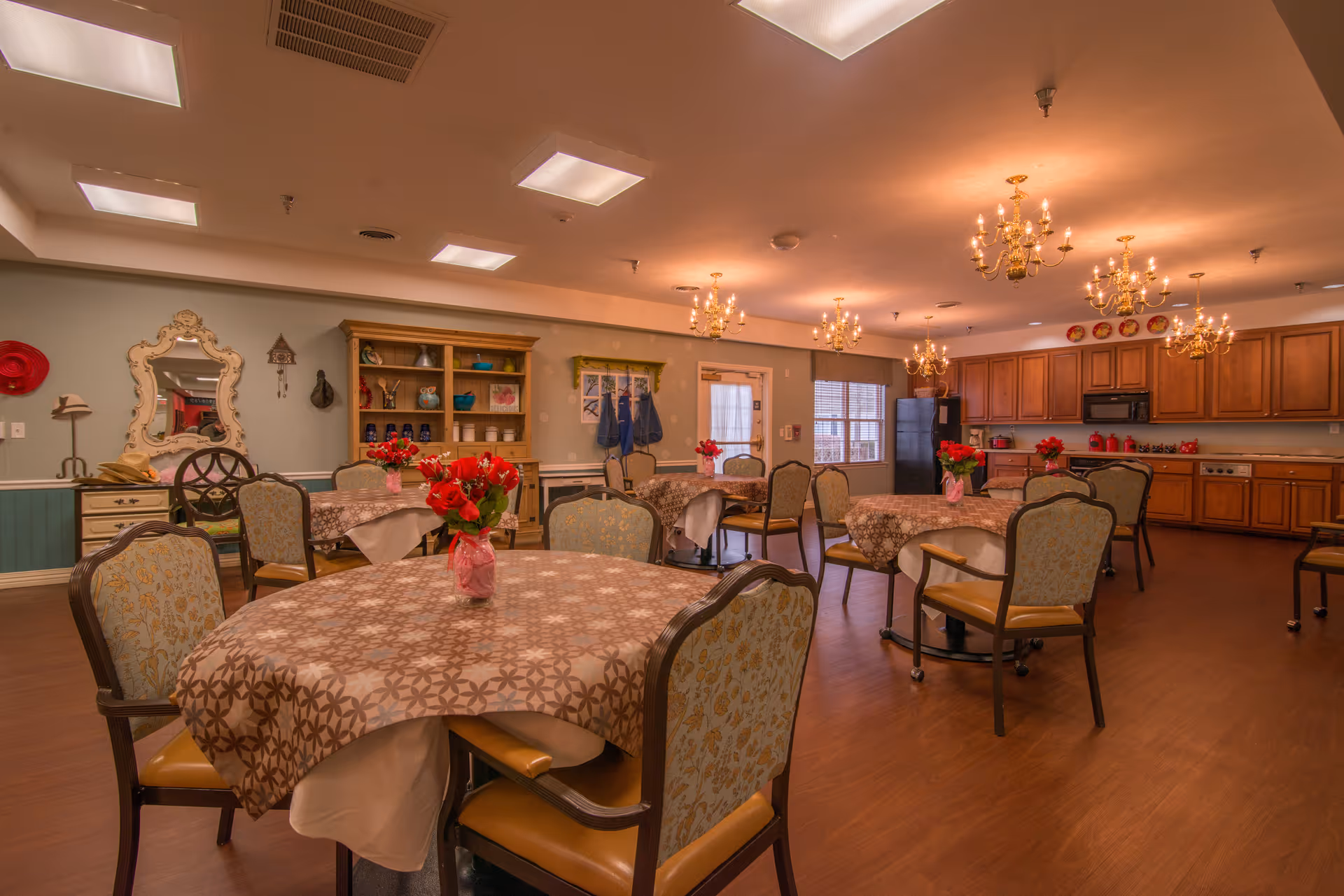 Warmly lit communal dining room with round tables covered in patterned tablecloths, floral centerpieces, upholstered chairs, chandeliers, and a kitchen area in the back.