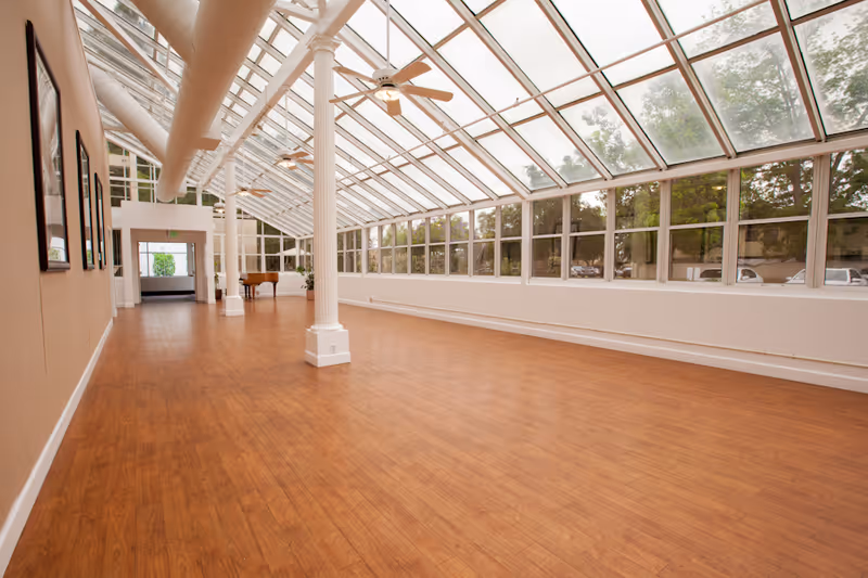 A spacious indoor room with a wooden floor and large glass windows and ceiling allowing natural light to fill the space. The room features white columns, ceiling fans, and a piano in the far corner. The walls have framed pictures and there is an exit door at the end of the room.