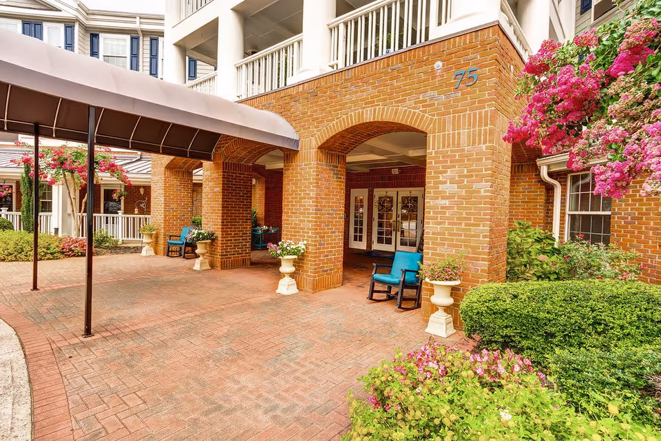 Entrance area of a senior living facility with brick arches, a covered walkway, potted plants, and blue rocking chairs. The building has white railings and flowering bushes nearby.