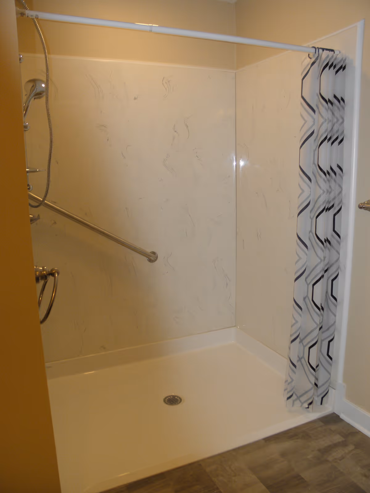 A walk-in shower with a handheld showerhead, a horizontal grab bar on the wall, a white shower base, and a shower curtain with a geometric pattern. The walls have a light marbled design and the floor outside the shower is wood-patterned.