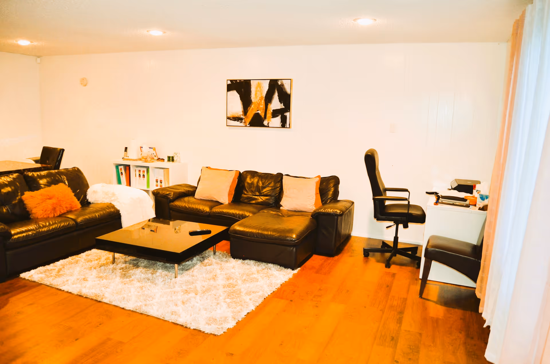 A cozy living room with two dark leather sofas adorned with cushions, a black coffee table on a fluffy white rug, a small white shelf with binders and items, a black office chair at a white desk with office supplies, and a modern abstract painting on the wall.