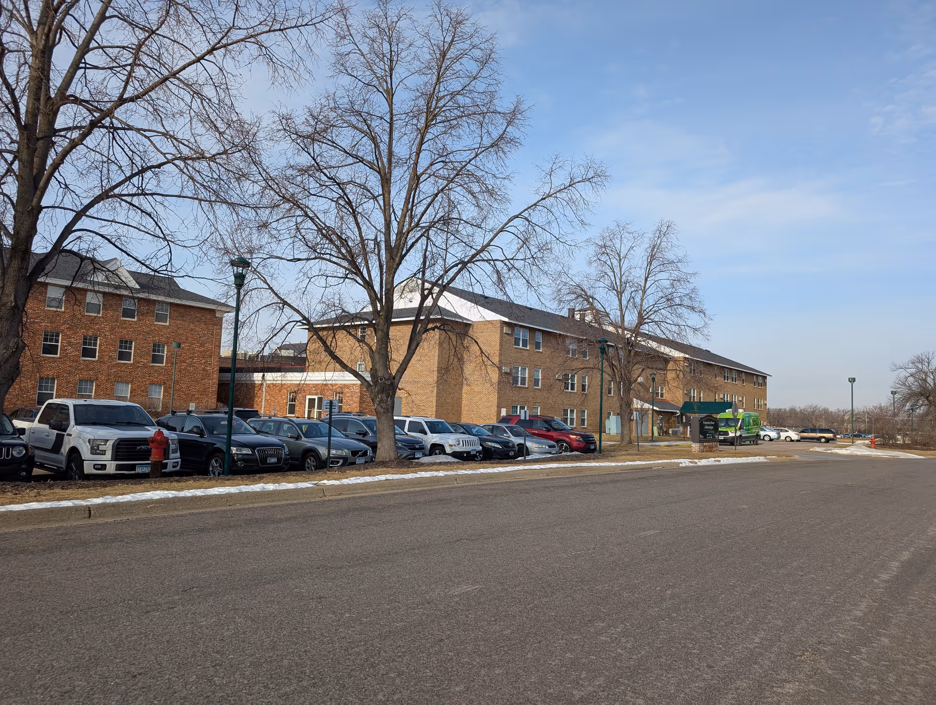 Exterior view of Newton Manor, a multi-story brick building with several windows. There are leafless trees and a row of parked cars along the street in front of the building. The sky is clear with some light clouds.