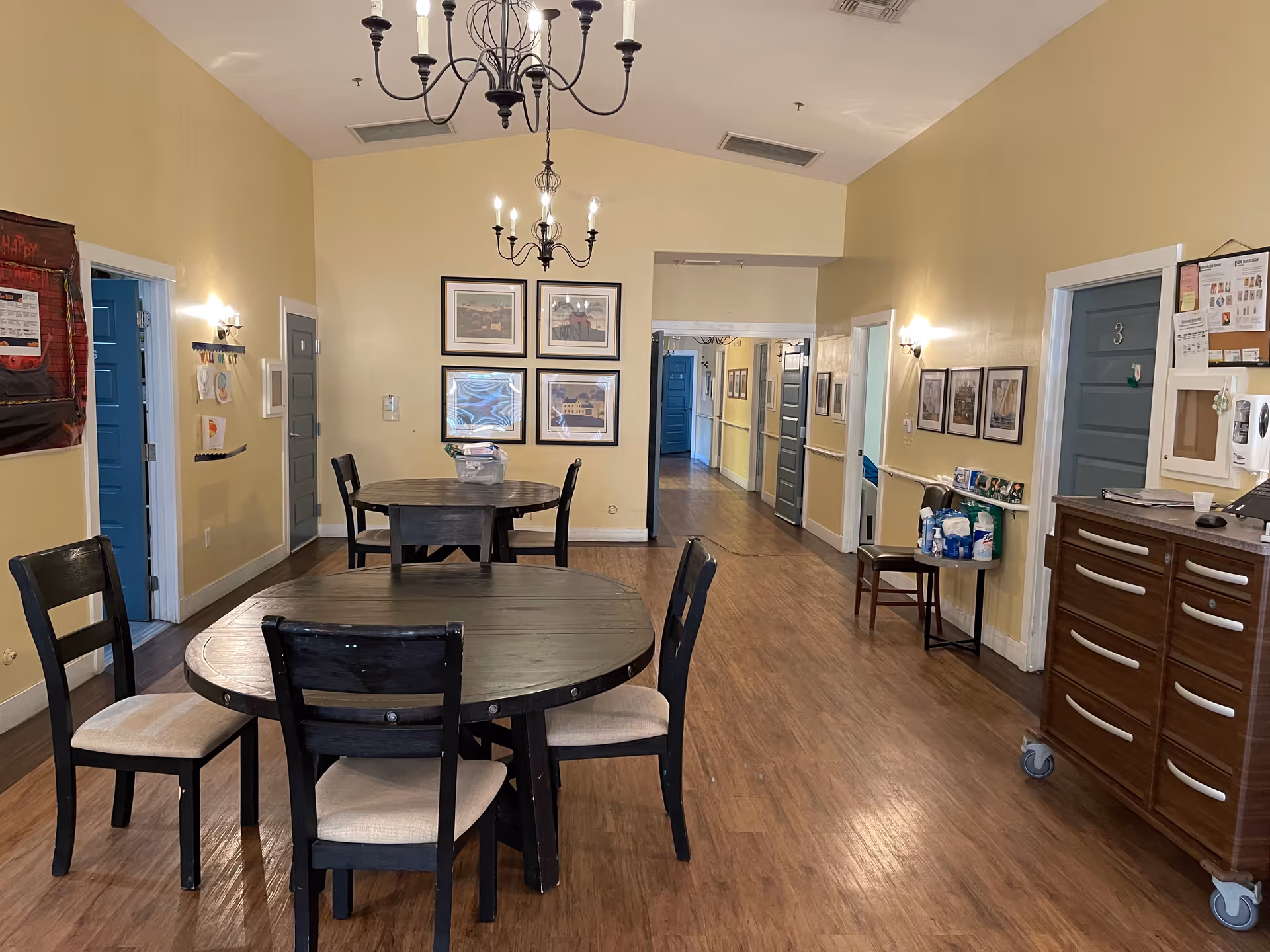 Interior view of a common area in an assisted living facility with two round wooden tables and chairs, yellow walls, wooden flooring, framed pictures on the walls, and a chandelier hanging from the ceiling. Several doors and a hallway are visible in the background, along with a wooden cabinet and a small table with supplies.