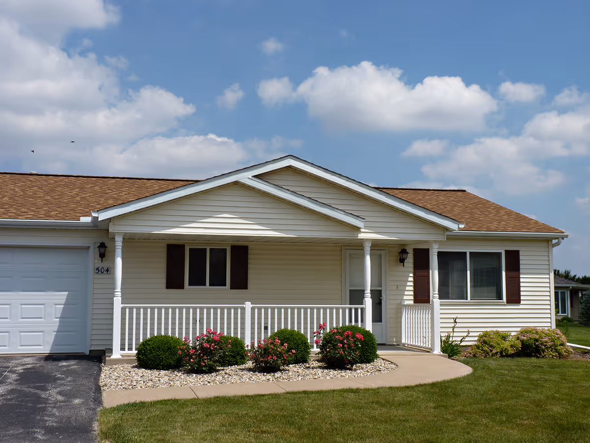 Single-story residential building with beige siding, brown roof shingles, and a small covered front porch with white railing. There are bushes and pink flowers planted in front of the porch, a concrete walkway leading to the front door, and a garage door on the left side. The sky is partly cloudy with blue patches.