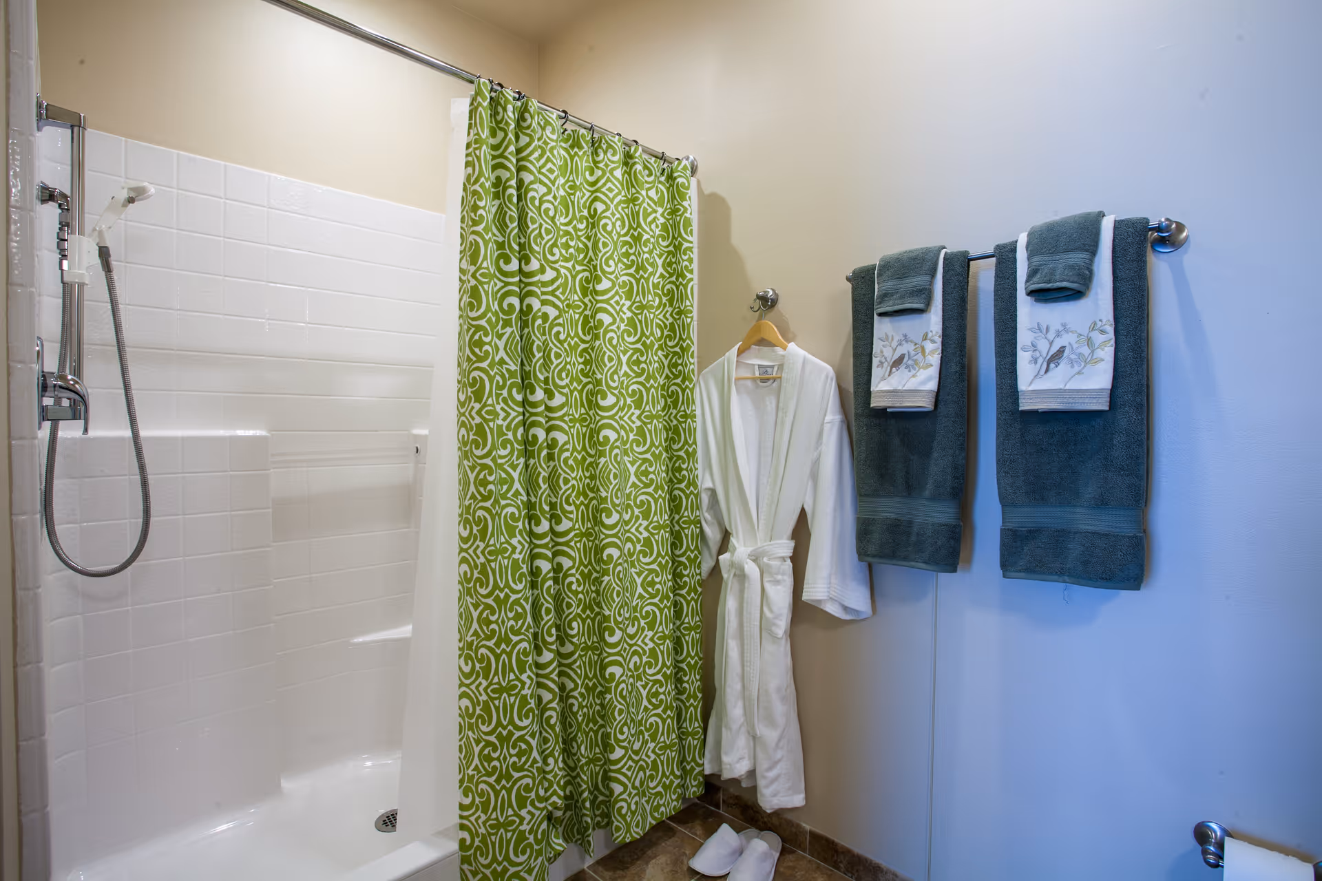 A bathroom shower area with white tiled walls and a green patterned shower curtain. Next to the shower, a white bathrobe is hanging on a hook, with white slippers on the floor beneath it. On the adjacent wall, two dark green towels with decorative white hand towels featuring bird and branch embroidery are neatly hung on a towel rack.