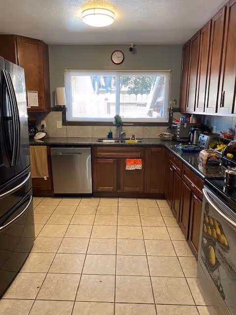 A kitchen with wooden cabinets, a tiled floor, and stainless steel appliances including a refrigerator, dishwasher, and oven. There is a window above the sink letting in natural light, and various kitchen items such as a coffee maker, toaster, and fruit bowl are on the countertops.
