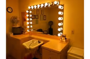 A well-lit vanity area with a large mirror surrounded by round light bulbs. Below the mirror is a countertop with a small white sink and various items on the surface. To the left, there is a small fan and a microwave on a cabinet. The walls are painted yellow, and several framed pictures or certificates are visible reflected in the mirror.