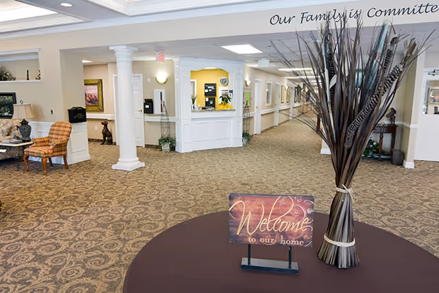 Reception area of a senior living facility with a 'Welcome' sign on a table, decorative vase, seating, columns, and a long carpeted hallway.