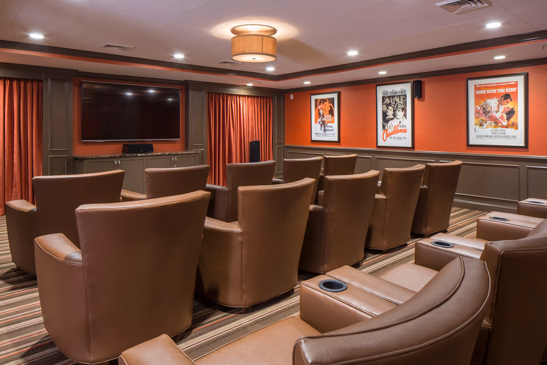 A cozy movie theater room with multiple rows of brown leather recliner chairs facing a large flat-screen TV mounted on a wall with red curtains on either side. The walls are painted orange with brown wainscoting, and three classic movie posters are displayed on the right wall. The ceiling has recessed lighting and a central light fixture.