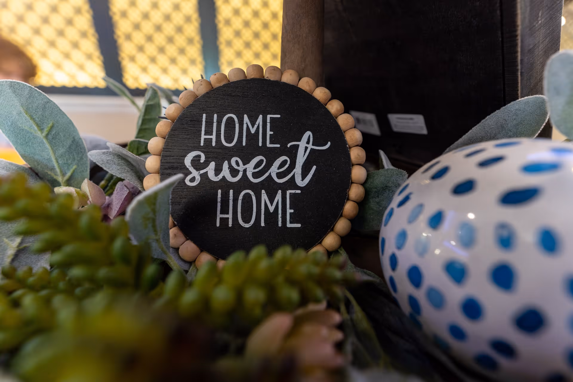 Close-up of a decorative sign with the words 'Home Sweet Home' surrounded by green foliage and a white ceramic object with blue spots.
