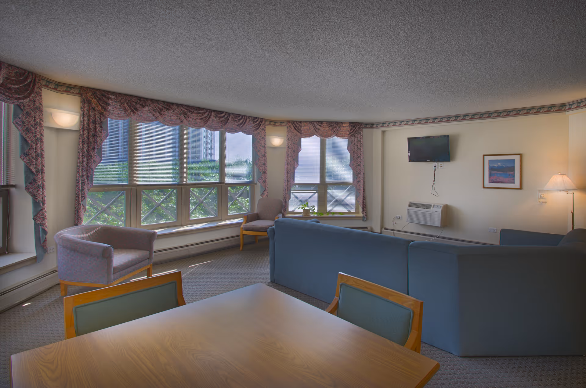 A cozy living room area in a senior living facility with large windows covered by patterned valances, two armchairs near the windows, a blue sectional sofa, a wall-mounted TV, a framed picture, and a floor lamp. A wooden table with chairs is in the foreground.