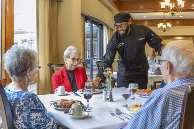 A waiter dressed in black serves food to three elderly people seated around a dining table in a well-lit dining room. The table is set with plates of food, glasses of red wine, and a small vase with flowers. The room has large windows with curtains and warm lighting fixtures on the ceiling.