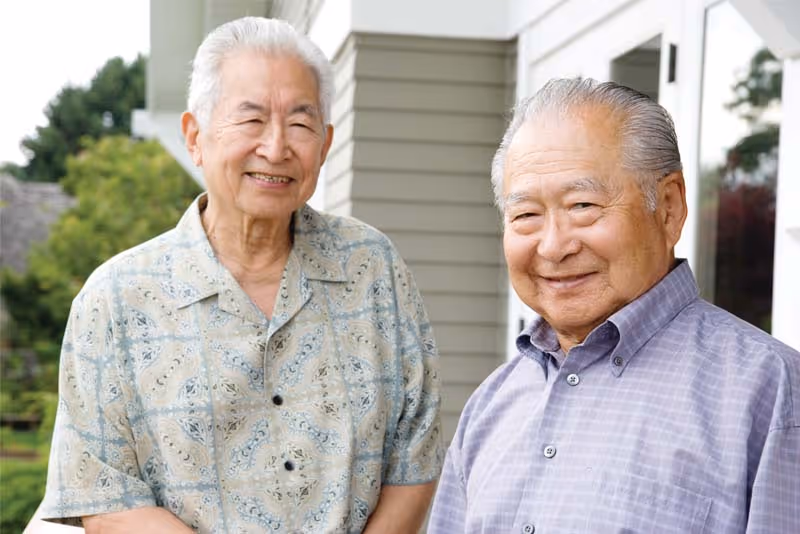 Two elderly men standing outside near a building with siding, smiling at the camera. There are trees and greenery in the background.