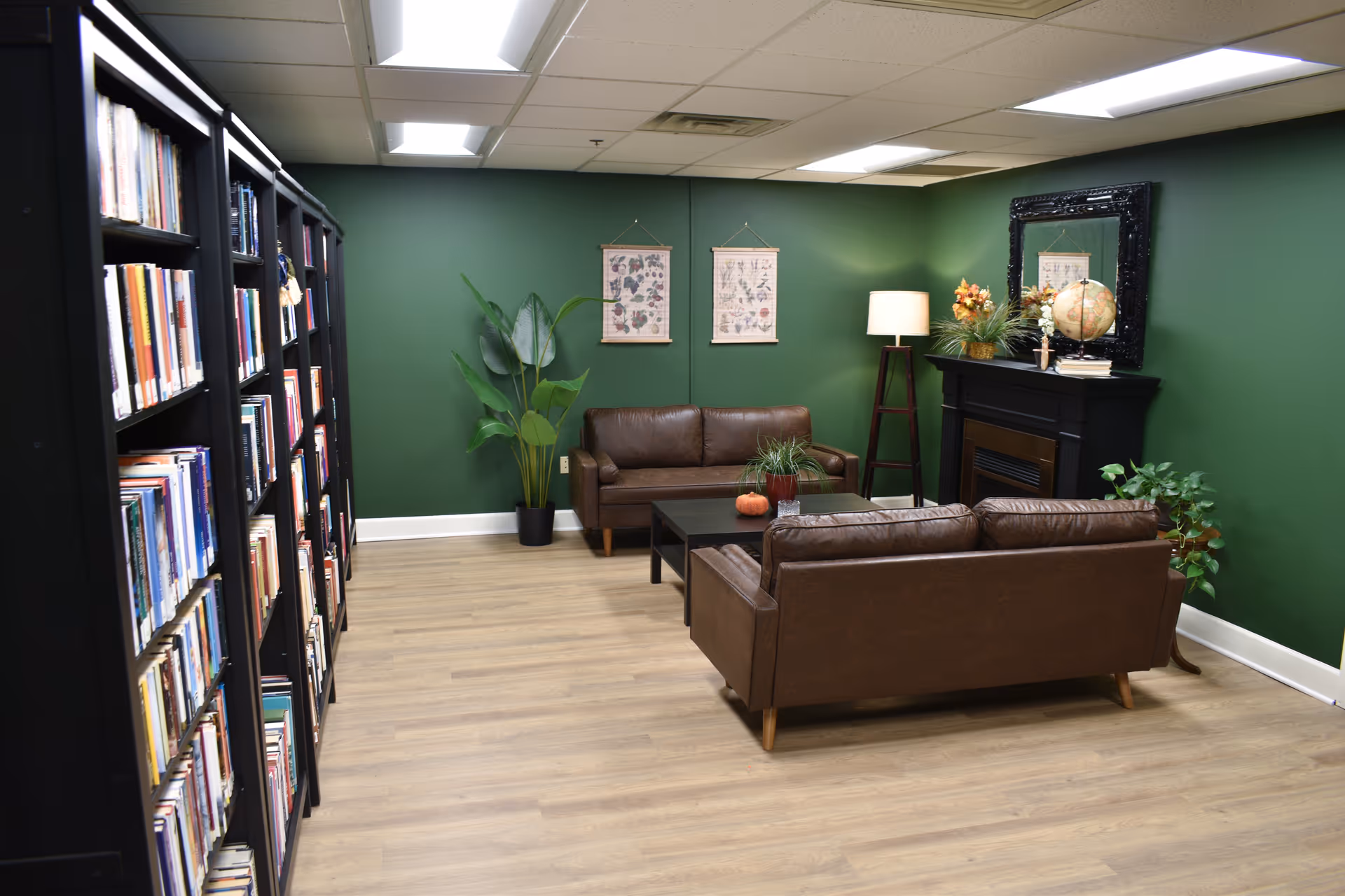 Cozy common room with brown leather sofas, bookshelves, green walls, plants, and a decorative fireplace.