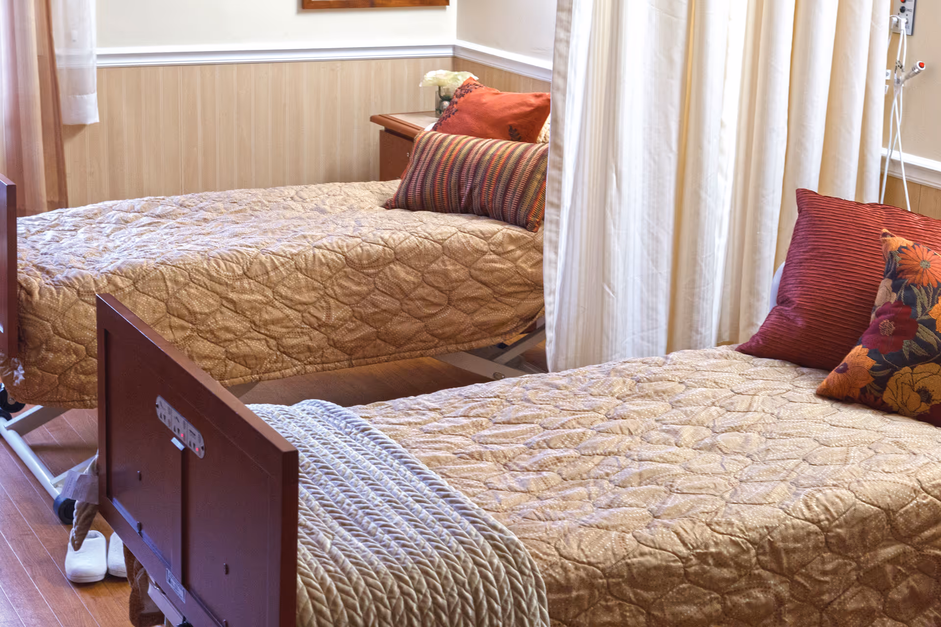 A nursing facility room with two hospital-style beds separated by a white privacy curtain. Each bed has a beige quilted bedspread and decorative pillows in red and floral patterns. There is a wooden nightstand with a flower vase between the beds, and the room has wood paneling on the lower half of the walls and hardwood flooring.