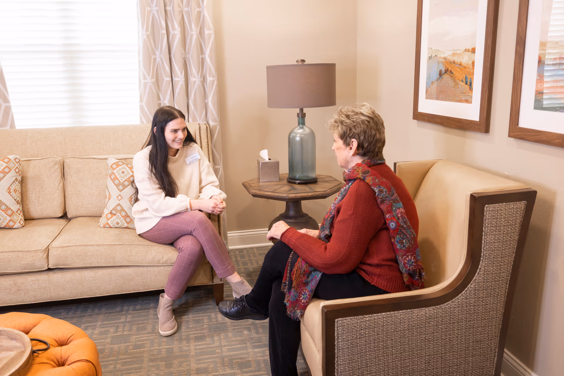 A young woman and an older woman sitting and talking in a cozy living room area with beige furniture, a side table with a lamp and tissue box, patterned curtains, and framed artwork on the wall.