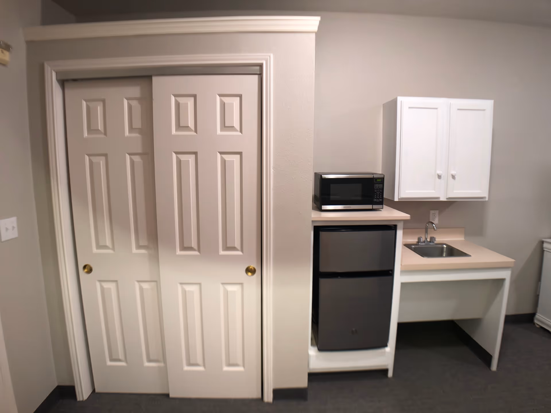 Small kitchenette area with a mini refrigerator, microwave on top, a sink with a faucet, and white cabinets mounted on the wall. To the left, there are white sliding closet doors with gold-colored knobs.